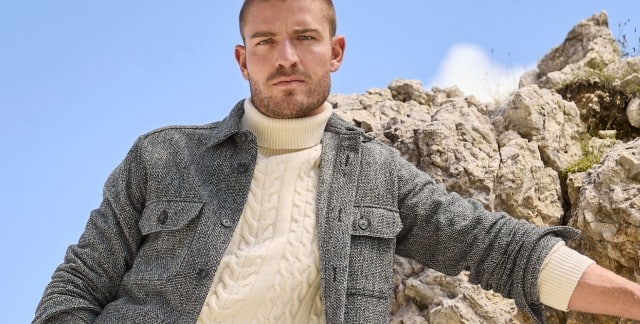 Ein Mann steht vor Felsen und trägt eine Hemd-Jacke in Schwarz/Weiß, darunter einen Rollkragen-Pullover mit Zopfmuster in Wollweiß.