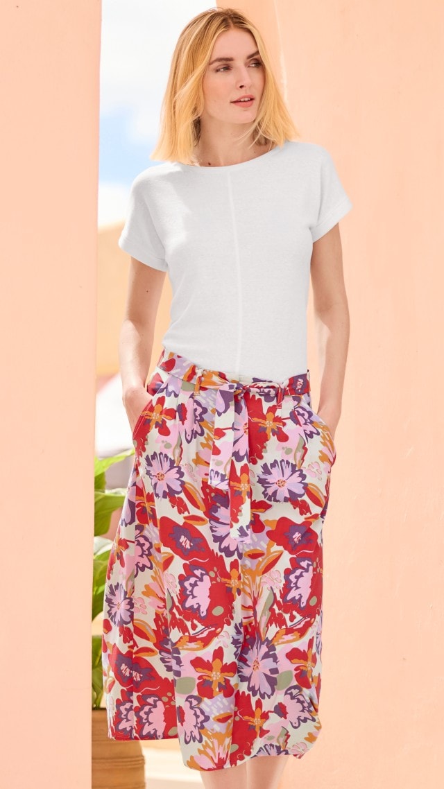 Eine Frau trägt ein schlichtes, weißes T-Shirt und einen farbenfrohen, knielangen Rock mit großem Blumenmuster in Rot-, Lila- und Gelbtönen. Ihr Stil wirkt sommerlich, frisch und modern.