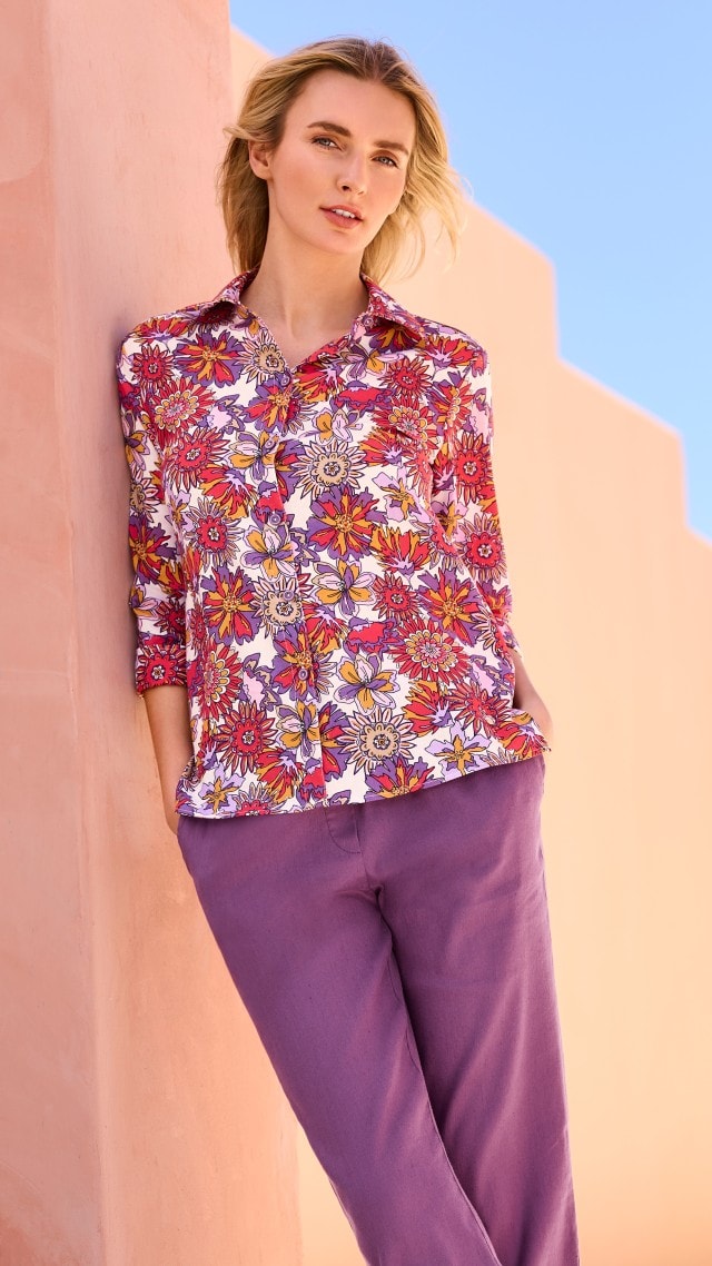 Eine Frau trägt eine bunte Bluse mit auffälligem Blumenmuster in Rot-, Lila- und Gelbtönen sowie eine lilafarbene Stoffhose. Ihr Stil ist modern, farbenfroh und sommerlich.