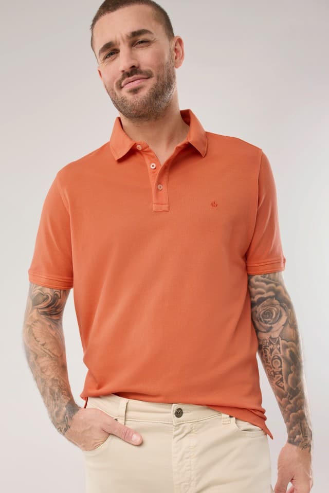Ein Mann trägt ein Polo-Shirt in Orange und hält seine Hand in der Hosentasche. Ein Mann trägt ein Polo-Shirt in Orange und hält seine Hand in der Hosentasche.