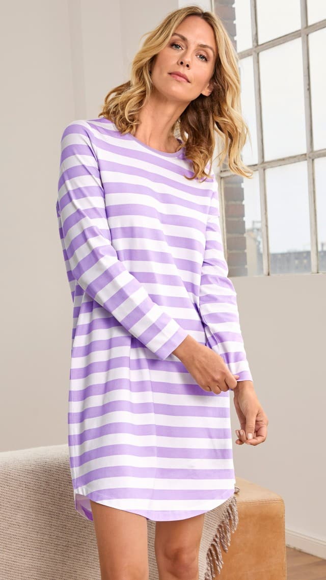 Eine Frau trägt ein locker fallendes, langärmeliges Kleid mit breiten lila‑weißen Streifen. Der Look wirkt frisch, leger und angenehm unkompliziert.