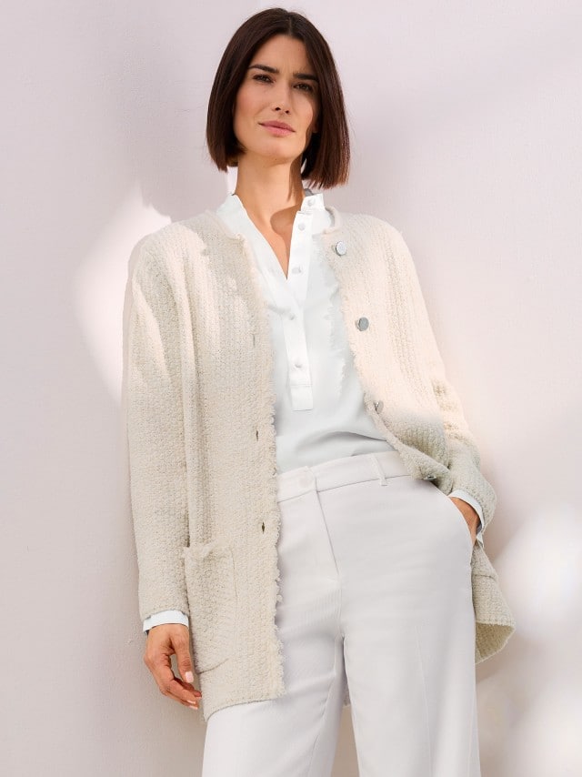 Frau trägt eine helle Strickjacke mit Knöpfen über einer weißen Bluse und weißen Stoffhosen – eleganter, monochromer Look in sanften Tönen.