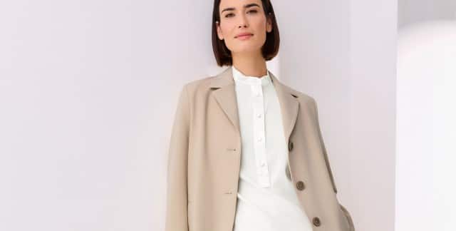 Frau in eleganter, beiger Jacke über einer weißen Bluse mit Stehkragen, steht vor weißem Hintergrund – schlichter und moderner Business-Look.