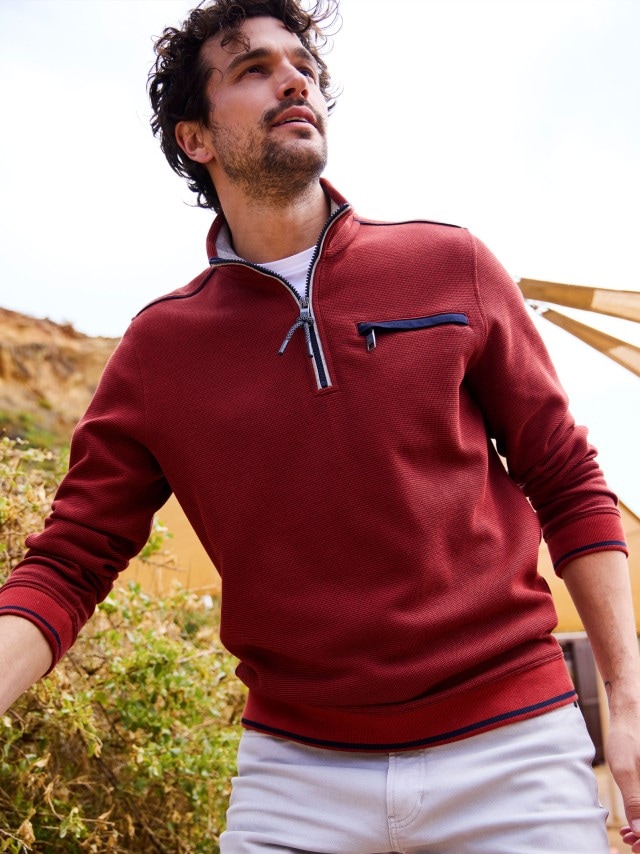 Ein Mann trägt einen roten Zipper-Pullover mit dunklen Details an Kragen und Brusttasche. Dazu kombiniert er eine helle Hose für einen sportlich-modernen Look.