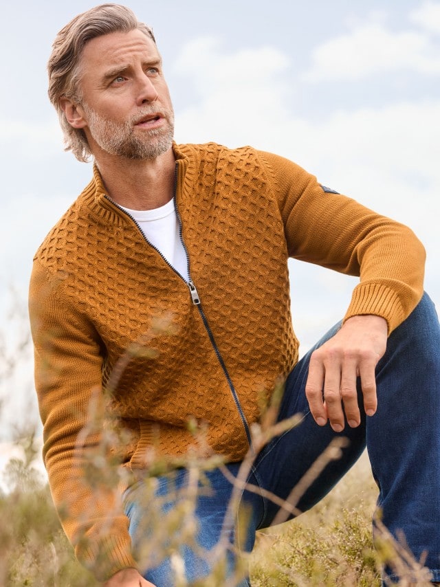 Ein Mann hockt auf einer Weide und trägt die Premium Baumwoll-Strickjacke in Messing, darunter ein weißes Shirt und dazu eine blaue Jeans.
