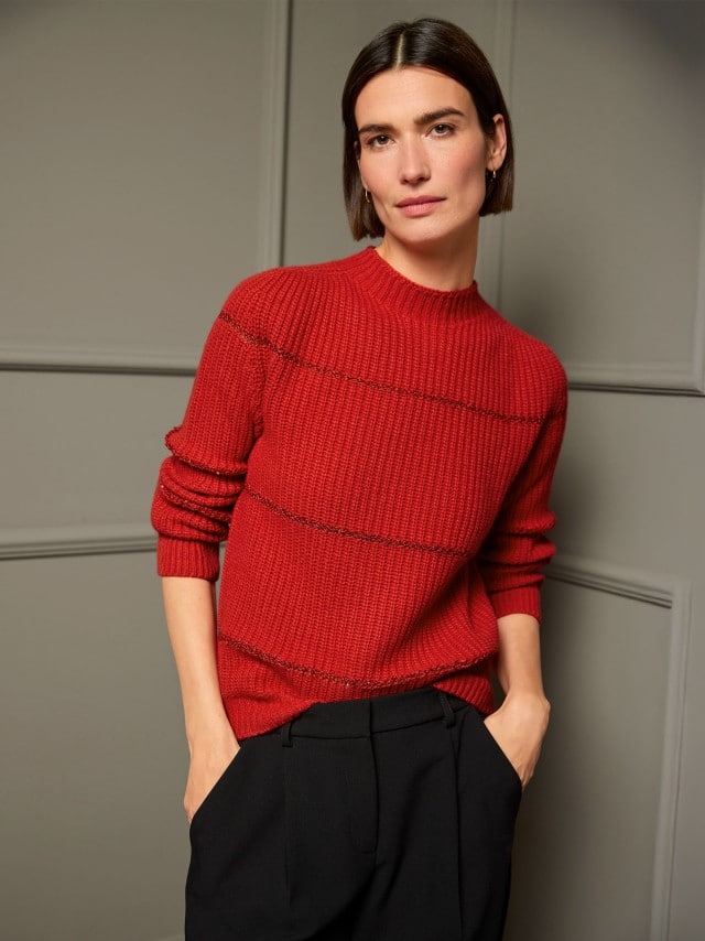 Eine Frau steht vor einer dunklen Wand und trägt einen roten Pullover mit Paillettenstreifen, dazu eine schwarze Hose. 