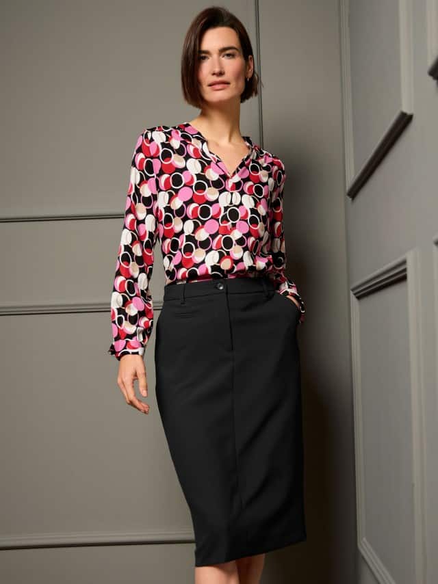 Eine Frau steht vor einer dunklen Wand und trägt eine bedruckte Easy Care Viskose Bluse, dazu eine schwarze Hose.