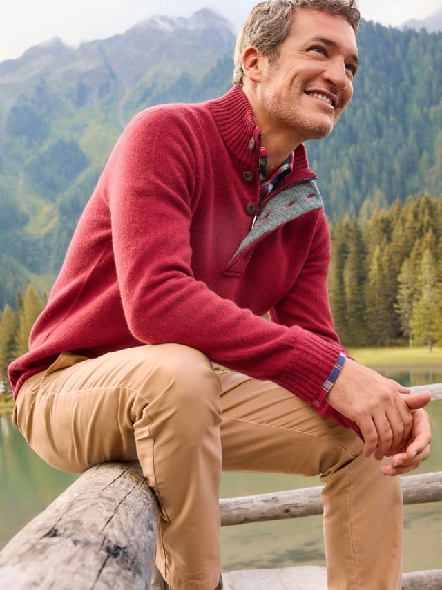 Ein Mann sitzt auf einem Holz-Geländer und trägt den Premium Lammwoll-Troyer in der Farbe Zinfandel, darunter eine kariertes Hemd und dazu eine Hose in Sandgelb.