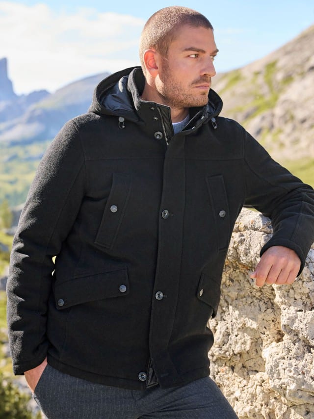 Ein Mann steht an einem Felsen und trägt eine schwarze Thermofleece Jacke, dazu eine Premium Sport-Flanell Hose in Dunkelgrau.