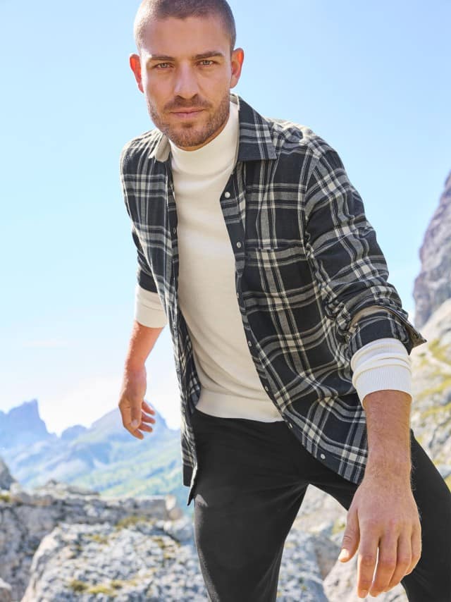 Ein Mann steht auf einem Felsen und trägt ein weißes Rollkragen-Shirt mit einem karierten Hemd darüber, dazu eine  schwarze Hose.