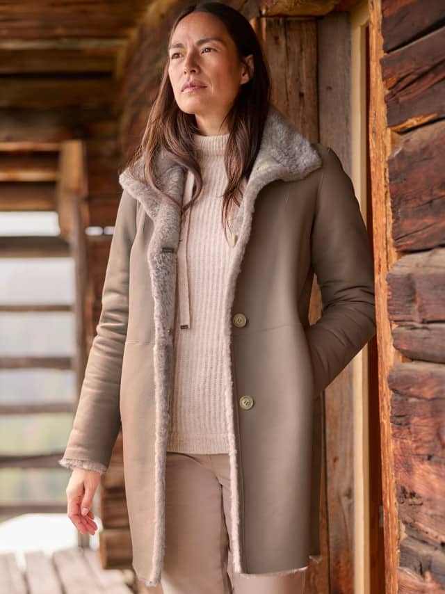 Eine Frau steht vor einer Holzhütte und trägt einen Rollkragen-Pullover, darüber den Premium Lammfell Wendemantel in Taupe, dazu eine Hose in Beige.