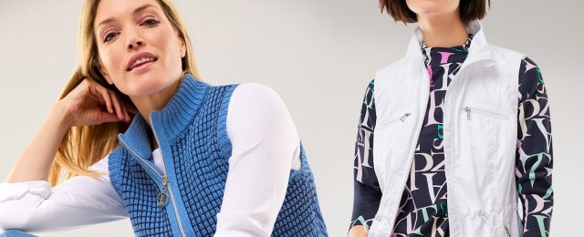 Zwei Frauen tragen moderne Westen: Eine kombiniert eine blaue Strickweste mit einem weißen Langarmshirt, die andere eine weiße, sportive Weste über einem gemusterten Shirt mit großen Buchstaben. Die Looks sind lässig und vielseitig für den Alltag geeignet.