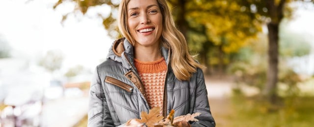 Eine Frau steht mit einer grauen Outdoor-Jacke in einem Park und hält Blätter in den Händen