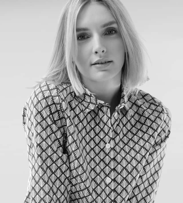 Ein blonde Frau trägt eine gemusterte Bluse.