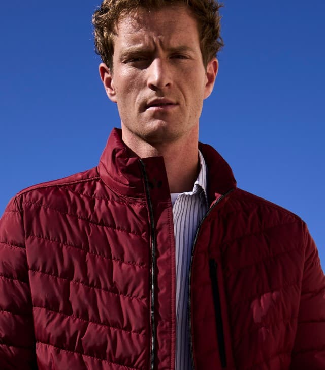 Ein Mann trägt eine gesteppte, weinrote Jacke über einem Hemd mit feinen Streifen. Sein Stil wirkt modern, frisch und passend für die Übergangszeit.