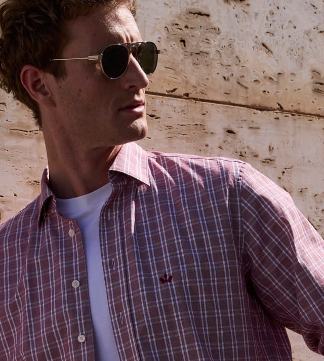 Ein Mann mit Sonnenbrille steht vor einer Natursteinwand und trägt ein rotes kariertes Hemd, darunter ein weißes Shirt.