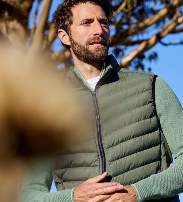 Ein Mann trägt eine gesteppte Weste in Grün, darunter einen passenden Pullover. Im Vordergrund sind gelbe Blüten zu sehen. 