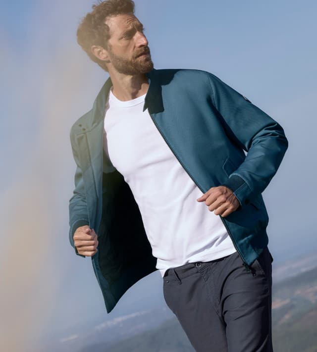Ein Mann steht auf einem Feld mit gelben Blumen und trägt einen Blouson in Blau, darunter ein weißes Shirt und dazu eine Hose in Marine.