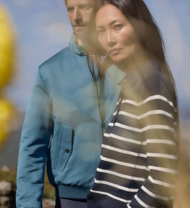 Ein Mann und eine Frau stehen auf einem Feld mit einzelnen Blumen. Er trägt einen blauen Blouson und sie einen gestreiften Pullover mit Kragen in Marine/Weiß. 