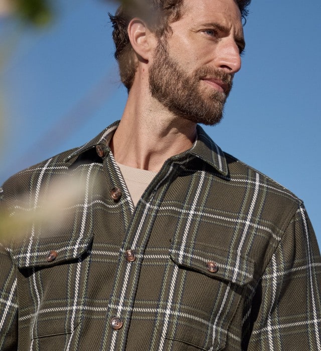 Ein Mann trägt ein Karo Overshirt in Oliv, darunter ein Shirt in Beige. Vor ihm ist eine gelbe Blume zu sehen.