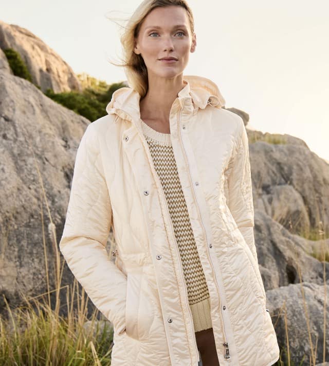 Eine Frau steht vor einer felsigen Landschaft und trägt eine helle Jacke über einem gestrickten Oberteil.