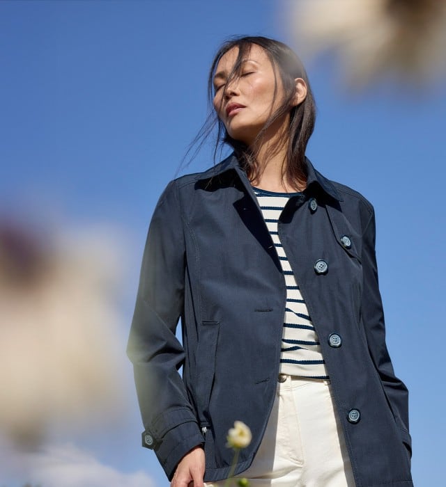 Eine Frau steht mit geschlossenen Augen auf einer Frühlingswiese und trägt eine Trenchjacke in Marine, darunter ein Baumwollshirt Ringelstruktur.