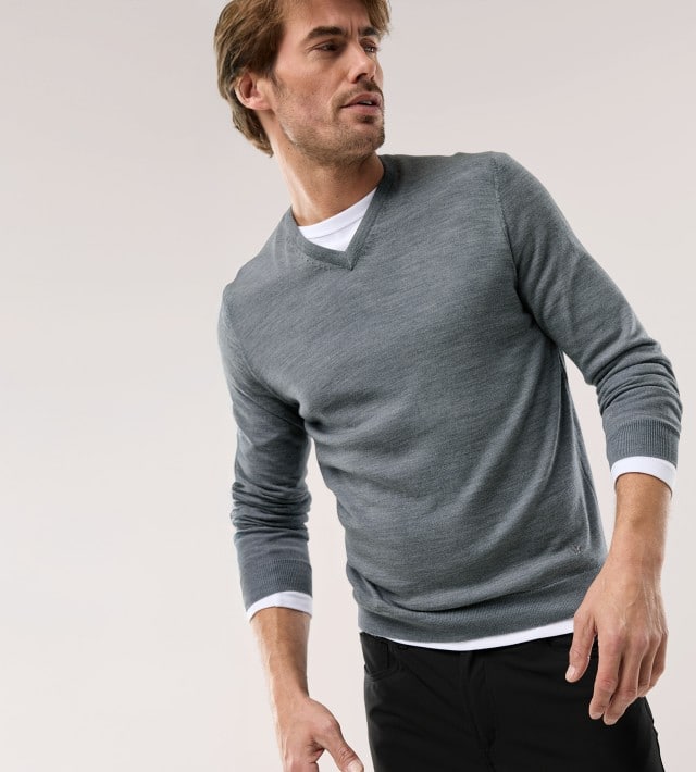Ein Mann vor einem hellen Hintergrund trägt einen grauen Pullover mit V-Ausschnitt, darunter ein weißes Shirt und dazu eine schwarze Hose.