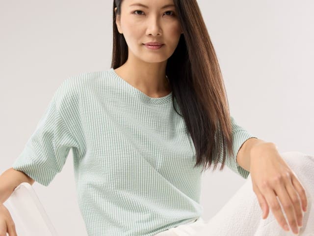 Eine Frau trägt ein Streifenshirt Seersucker in der Farbe Salbei, dazu eine weiße Hose.