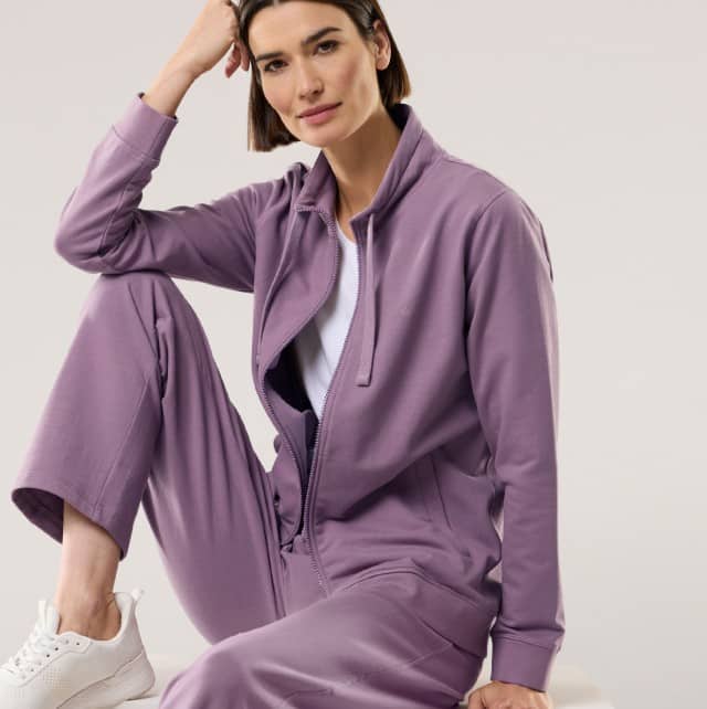Eine Frau sitzt auf dem Boden und trägt eine Sweat Jacke in der Farbe Mauve, mit passender Hose.