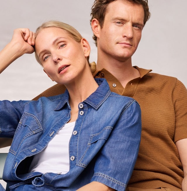 Ein Frau trägt ein Jeanshemd, darunter ein weißes Shirt und dazu eine passende Hose. Ein Mann in einem Strickpolo in der Farbe Latte. 
