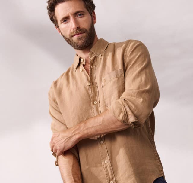 Ein Mann trägt ein Leinenhemd in Beige mit aufgekrempelten Ärmeln, dazu eine dunkle Hose.