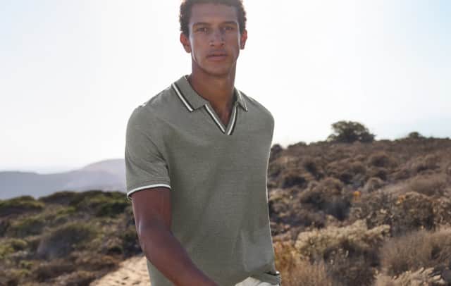 Ein Mann steht auf einem Hügel und trägt ein Polo-Shirt in Oliv, dazu eine weiße Hose.