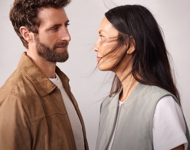 Ein Mann, in einem Leinen-Hemd in der Farbe Latte, steht neben einer Frau, die eine Jacquardweste in Khaki trägt, mit einem weißen Shirt darunter. 