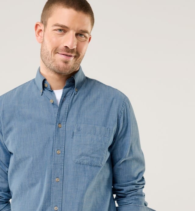 Ein Mann trägt ein hellblaues Jeanshemd über einem weißen T-Shirt und lächelt leicht. Der Stil wirkt locker und modern.