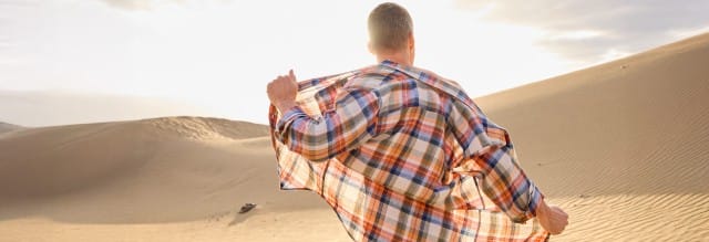Ein Mann steht in einer Wüstenlandschaft und trägt ein farbenfrohes kariertes Hemd im Freizeitstil, das er im Wind ausbreitet. Die Sonne scheint hell im Hintergrund.