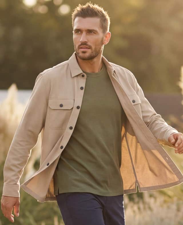 Ein Mann steht zwischen Gräsern und trägt eine helle Hemdjacke, darunter ein Shirt in Khaki. 