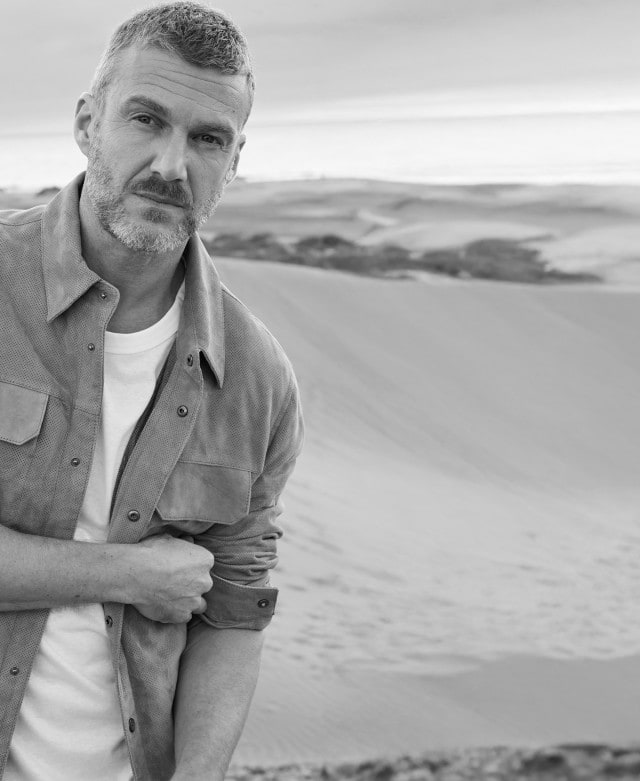 Ein Mann steht in einer Dünenlandschaft und trägt ein helles T-Shirt unter einer offenen Hemdjacke mit Brusttaschen.