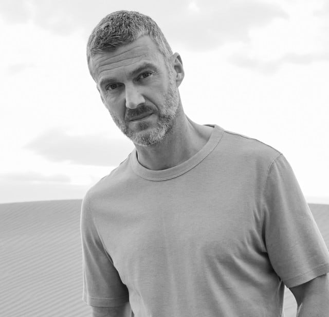 Ein Mann steht in den Dünen und trägt ein T-Shirt.