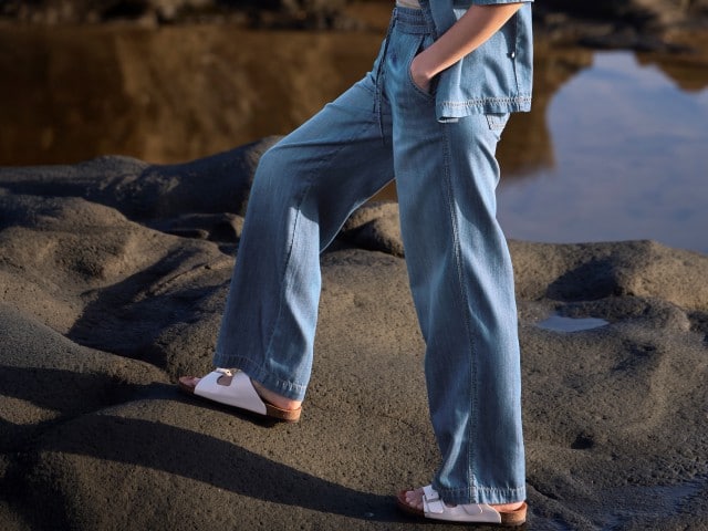 Eine Person trägt einen blauen Denim-Zweiteiler aus kurzärmliger Jacke und weit geschnittener Hose sowie weiße Riemchensandalen – ein lässiger Sommerlook.
