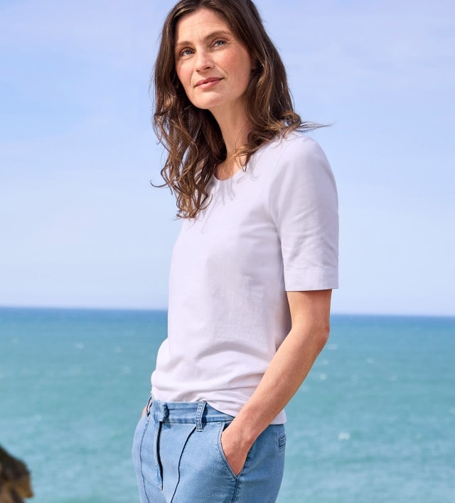 Eine Frau trägt ein schlichtes weißes T-Shirt und eine hellblaue Jeans. Ihr Stil ist entspannt und zeitlos, ideal für einen Tag am Meer.