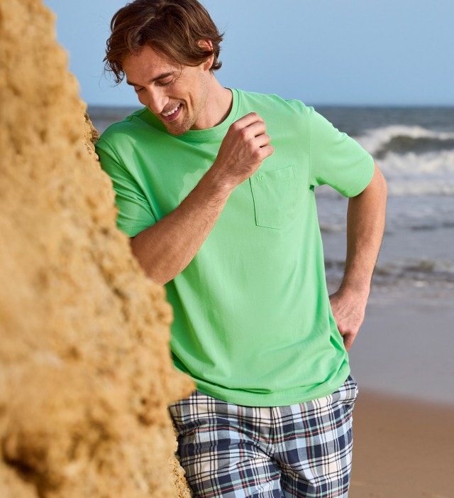Ein Mann steht an einem Felsen am Meer und trägt ein hellgrünes T-Shirt mit aufgesetzter Tasche auf der Brust.