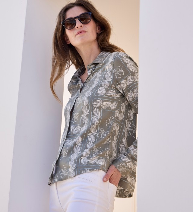 Eine Frau mit Sonnenbrille trägt eine gemusterte Bluse, dazu eine weiße Hose.