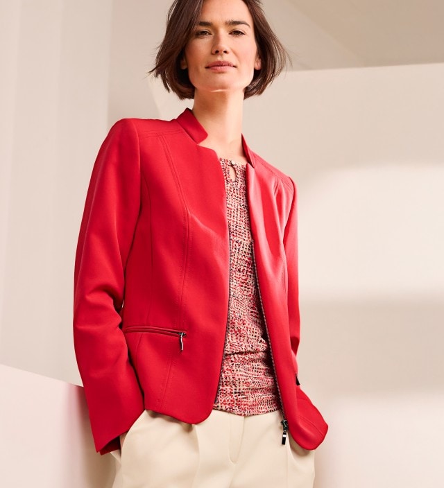 Eine Frau trägt einen roten Blazer mit Reißverschluss, darunter ein gemustertes Shirt und dazu eine helle Hose.