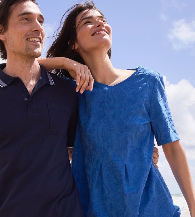 Ein Mann in einem dunkelblauen Polo-Shirt und eine Frau in einem blauen Kleid mit Stickereien, stehen am Strand.