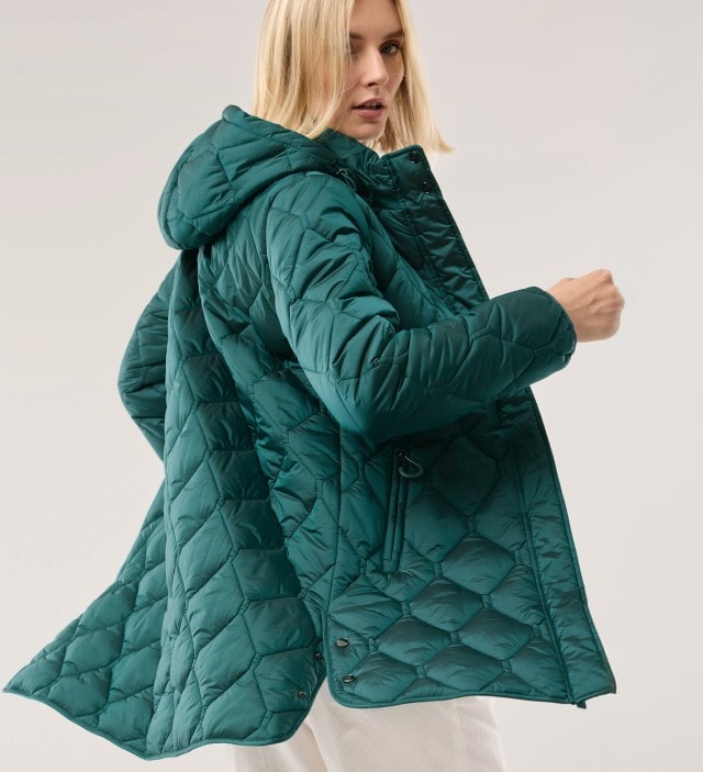 Frau in schwungvoller Bewegung trägt eine dunkelgrüne Steppjacke und helle Hose vor einem neutralen Hintergrund und schaut in die Kamera.