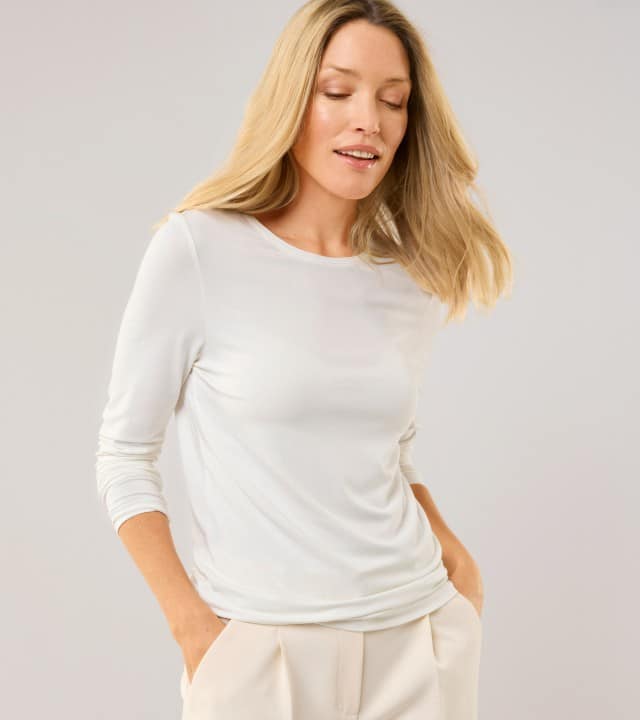 Eine Frau schaut nach unten und trägt ein weißes Langarm-Shirt, dazu eine Hose in Creme.