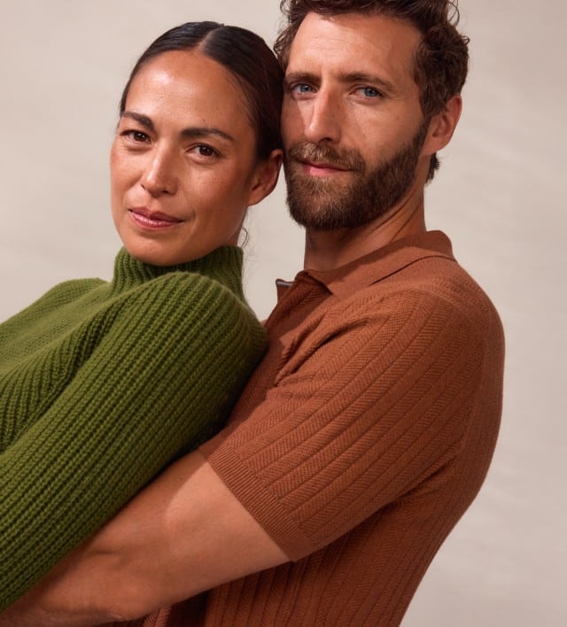 Ein Mann trägt ein kurzärmeliges, fein geripptes Oberteil in einem warmen Rostbraun und umarmt eine Frau in einem grob gestrickten, olivgrünen Pullover mit hohem Kragen.