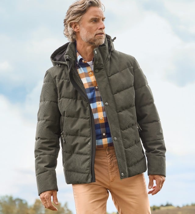 Ein Mann trägt eine olivgrüne Steppjacke über einem karierten Hemd in Blau-, Weiß- und Ockertönen sowie beigefarbene Chinos; der Look wirkt modern und herbstlich.