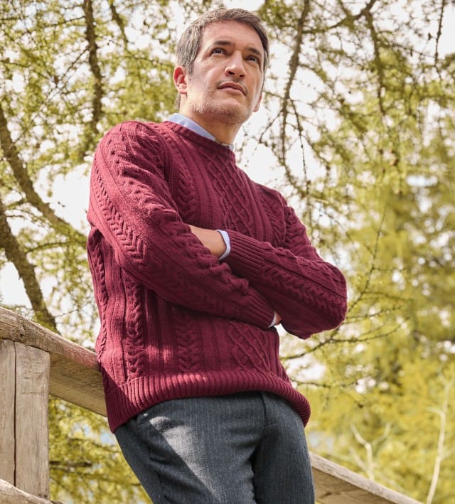 Ein Mann steht vor einem Baum und trägt den Aran-Pullover in Bordeaux, darunter ein hellblaues Hemd und dazu eine Sport-Flanell Hose in Anthrazit.