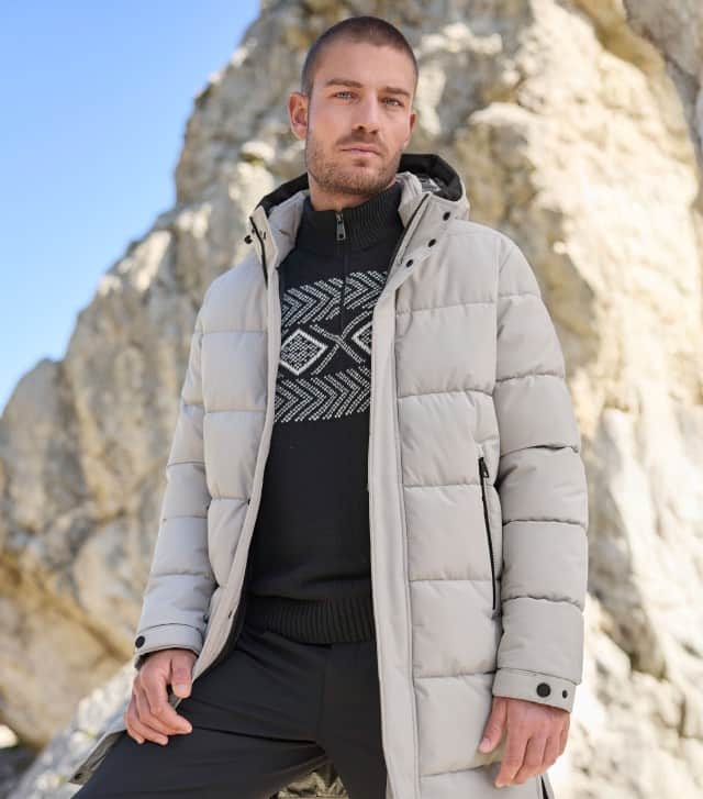 Ein Mann steht vor einer Felswand und trägt einen Thermo Steppmantel in Ecru, darunter einen Norweger Troyer in Schwarz und eine dunkele Hose.